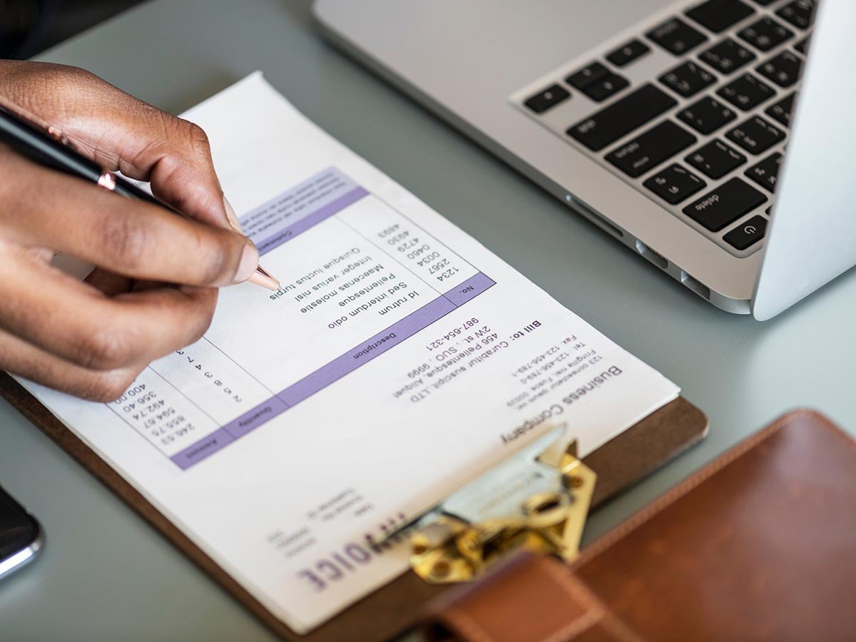 Hand holding a pen over an invoice on the desk next to a laptop